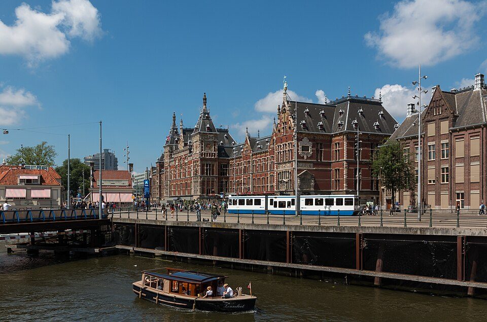 Fachada de la Estación Central de Ámsterdam bajo un cielo azul, con un tranvía pasando por delante junto al agua