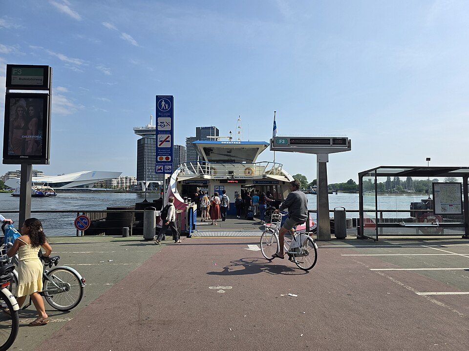 Cyclists and pedestrians at the free GVB ferry landing behind Amsterdam Central Station, with Amsterdam Noord across the IJ