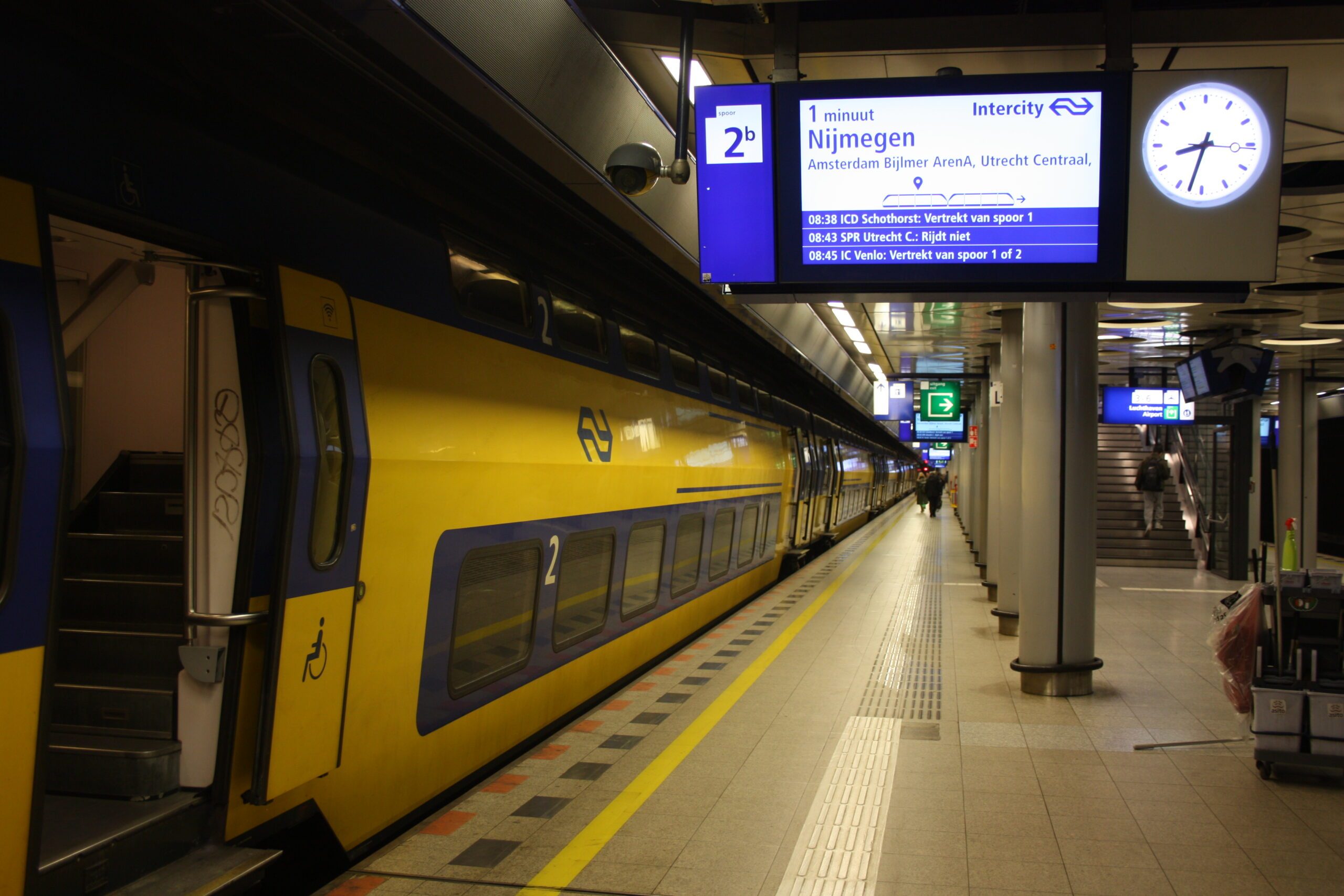 Gele dubbeldekker van NS aan een ondergronds perron van treinstation Schiphol Airport, met vertrekbord en klok